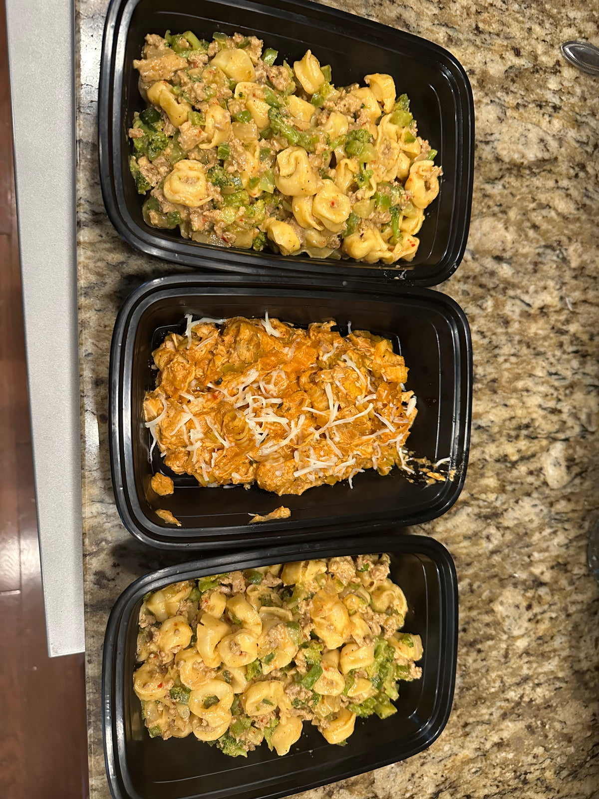 Three open meal-prep containers with pasta dishes on a kitchen countertop.
